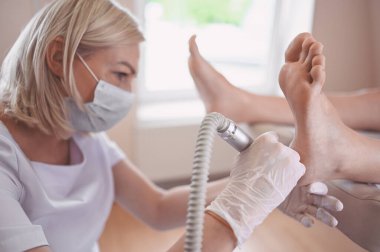Ayak bileme ekipmanları kullanan ve ayak pedikürü için işlem cilası yapan pediatrist doktor. Beyaz eldivenli Podoloji Güzellik Uzmanı müşteri bacaklarını nasırdan ve mısırdan profesyonel aletlerle temizliyor.