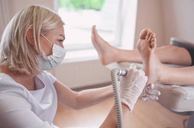 Ayak bileme ekipmanı kullanan ve ayak pedikürü için işlem cilası yapan bir pediatrist. Beyaz eldivenli Podoloji Güzellik Uzmanı müşteri bacaklarını nasırdan ve mısırdan profesyonel aletlerle temizliyor.