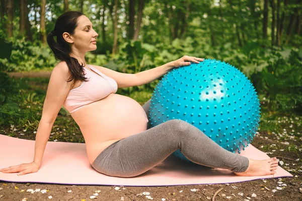 Beautiful young pregnant woman doing exercising with fitness pilates blue ball in park outdoor. Sitting and relaxing on pink yoga mat. Active future mother sport lifestyle. Healthy pregnancy concept.