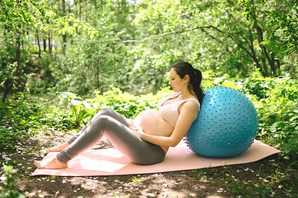 漂亮的年轻孕妇带着健身枕头在户外公园里做运动 坐在粉色瑜伽垫上休息 积极的未来母亲运动的生活方式 健康怀孕概念 — 图库照片
