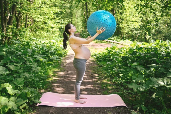 美丽的年轻孕妇在户外公园里做瑜伽运动 带着健身普拉提球 用双手站立和握住 积极的未来母亲运动的生活方式 健康怀孕概念 — 图库照片