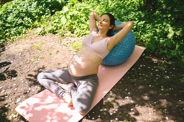 漂亮的年轻孕妇带着健身枕头在户外公园里做运动 坐在粉色瑜伽垫上休息 积极的未来母亲运动的生活方式 健康怀孕概念 — 图库照片