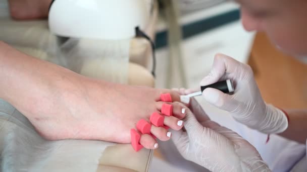 Pedicurist κάνει λευκό βερνίκι νυχιών στα πόδια του πελάτη χρησιμοποιώντας shellac λάμπα και το δάχτυλο του ποδιού διαχωριστή. Επαγγελματική ιατρική διαδικασία πεντικιούρ. Θεραπεία ποδιών στο σαλόνι SPA. Κλινική Ποντιατρία. Χέρια σε γάντια. — Αρχείο Βίντεο