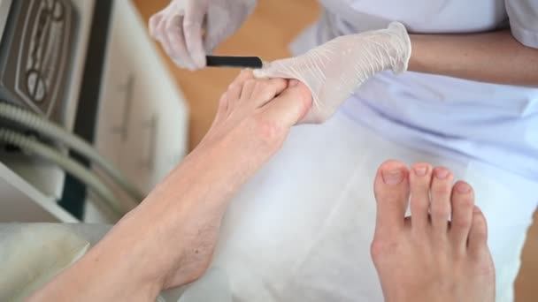 Pedicurist doing professional medical pedicure procedure in beauty salon using nail file. Foot treatment in SPA salon. Podiatry clinic. Beautician doctor hands in white gloves with female legs — Stock Video
