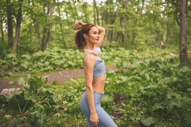 Mavi spor kıyafetleri içinde genç, güzel, sıska bir kız büyük beyaz kulaklıklarla müzik dinliyor dışarıda yaz parkında spor ve spor yapıyor. Mükemmel vücutlu bir kız yoga ya da egzersiz yapıyor.