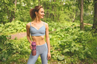 Mavi spor kıyafetli genç, güzel, sıska kız dışarıda yaz parkında spor ve spor yaparken bir şişeden su içiyor. Mükemmel vücutlu bir kız yoga ya da egzersiz yapıyor.