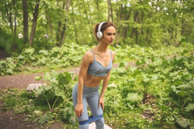 Mavi spor giyimli, genç, güzel, sıska, güzel bir kız. Büyük, beyaz kulaklıklı. Açık yeşil parkta spor salonunda spor yaparken spor yapıyor. Mükemmel vücutlu kız, spor yogası.