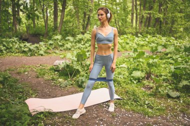 Mavi spor kıyafetli, büyük beyaz kulaklıklı, spor kıyafetli genç ve güzel bir kız yaz yeşili parkta pembe yoga minderiyle spor yaparken spor yapıyor. Mükemmel vücutlu kız.