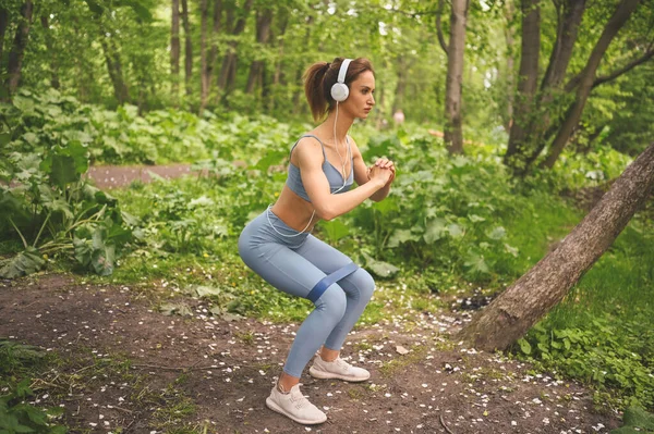 穿着蓝色运动服的年轻 体形苗条的女孩 戴着大大的白色耳机 在室外的夏季绿色公园里做着附有健身训练口香糖的蹲式锻炼 完美身材的女孩 运动瑜伽 — 图库照片
