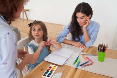 Gülümseyen anne ve kız ders için hazırlanıyor ve masada kalemler ve boyalarla resim çiziyor. Anaokulunun ebeveyni ve öğrencisiyim. Sonbaharın ilk günü. İlkokuldaki kız, okula geri döndü..