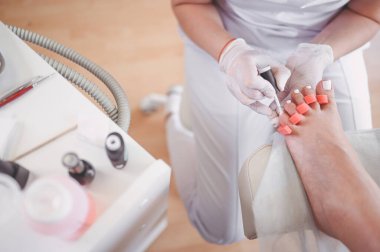 Pedikürcü, ayak parmağı ayırıcı kullanarak hasta bacaklarına beyaz oje sürüyor. Profesyonel tıbbi pedikür prosedürü. SPA salonunda ayak tedavisi. Ayak hastalıkları kliniği. Beyaz eldivenli güzellik uzmanı elleri