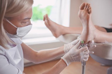 Ayak bileme ekipmanları kullanan ve ayak pedikürü için işlem cilası yapan pediatrist doktor. Beyaz eldivenli Podoloji Güzellik Uzmanı müşteri bacaklarını nasırdan ve mısırdan profesyonel aletlerle temizliyor.