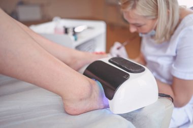 Pedikürcü, beyaz tırnak cilası sürüyor. Profesyonel tıbbi pedikür prosedürü. SPA salonunda ayak tedavisi. Ayak hastalıkları kliniği. Güzellik uzmanı beyaz eldivenlerle ellerini uzattı.