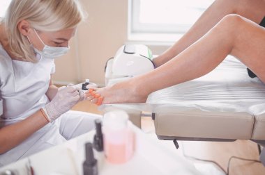 Pedikürcü, beyaz tırnak cilası sürüyor. Profesyonel tıbbi pedikür prosedürü. SPA salonunda ayak tedavisi. Ayak hastalıkları kliniği. Güzellik uzmanı beyaz eldivenlerle ellerini uzattı.