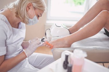 Pedikürcü, beyaz tırnak cilası sürüyor. Profesyonel tıbbi pedikür prosedürü. SPA salonunda ayak tedavisi. Ayak hastalıkları kliniği. Güzellik uzmanı beyaz eldivenlerle ellerini uzattı.