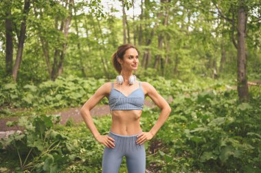 Mavi spor kıyafetleri içinde genç, güzel, sıska bir kız büyük beyaz kulaklıklarla müzik dinliyor dışarıda yaz parkında spor ve spor yapıyor. Mükemmel vücutlu bir kız yoga ya da egzersiz yapıyor.