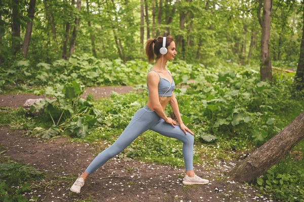 穿着蓝色运动服的年轻 体形苗条的女孩 戴着大大的白色耳机 在室外的夏季公园里做运动和健身 完美身材的女孩做瑜伽或运动 — 图库照片