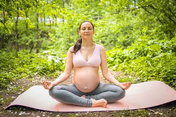 Hermosa joven embarazada haciendo ejercicio de yoga en el parque al aire libre. Sentarse y relajarse en la esterilla de yoga rosa. Estilo de vida activo futuro deporte madre. Concepto de embarazo saludable — Foto de Stock