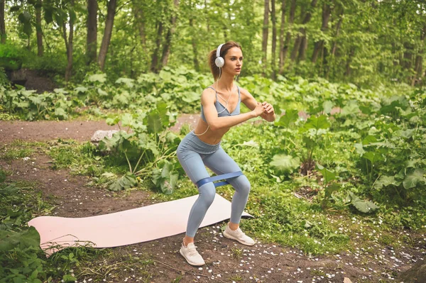 身穿蓝色运动服，身穿白色大耳机的年轻漂亮苗条身材的女孩，在室外的夏季绿地公园里，用粉色瑜伽垫做着附有健身训练口香糖的蹲式练习。身材完美的女孩. — 图库照片