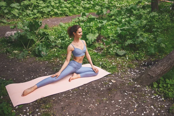 Hermosa mujer en forma haciendo estiramiento de gimnasia dividida con esterilla de yoga, ropa deportiva. Musculosa chica delgada atractiva con vientre plano en el parque de verano al aire libre. Cuidado corporal, saludable, fitness, concepto de vida deportiva. —  Fotos de Stock