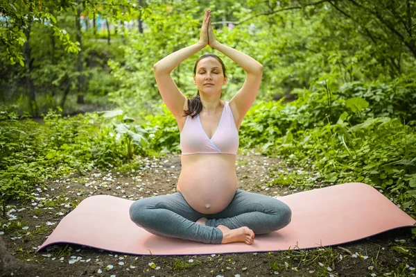 Красива молода вагітна жінка займається вправами йоги в парку на відкритому повітрі. Сидячи і розслабляючись на рожевому килимку йоги. Активний майбутній маминий спортивний спосіб життя. Концепція здорової вагітності — стокове фото