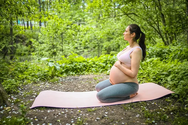 Beautiful young pregnant woman doing yoga exercising in park outdoor. Sitting and relaxing on pink yoga mat. Active future mother sport lifestyle. Healthy pregnancy concept