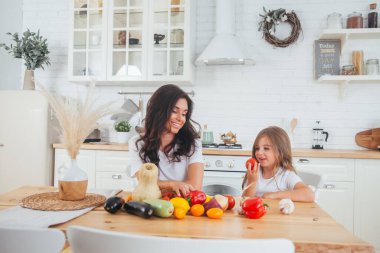 Evde poz veren mutlu bir anne ve çocuk. Güzel bir anne ve küçük kız İskandinav tarzı bir iç mekanda, beyaz bir mutfakta salata için sebze hazırlıyor. Sağlıklı yemek.