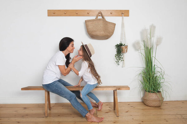 Mother puts on her little daughter a straw hat sitting on a bench in the Scandinavian style interior. Happy loving family having fun at home going for a walk