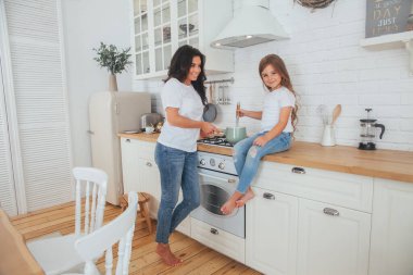 Mutlu bir aile yemeği hazırlıyor. Gülümseyen anne ve kız çocuğu mutfakta yemek pişiriyor ve eğleniyorlar. Ev yapımı sağlıklı yiyecekler. Beyaz İskandinav tarzı iç mekanda küçük bir yardımcı
