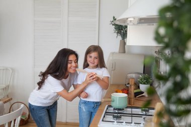 Mutlu bir aile yemeği hazırlıyor. Gülümseyen anne ve kız çocuğu mutfakta yemek pişiriyor ve eğleniyorlar. Ev yapımı sağlıklı yiyecekler. Beyaz İskandinav tarzı iç mekanda küçük bir yardımcı