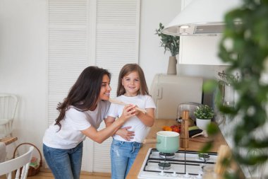 Mutlu bir aile yemeği hazırlıyor. Gülümseyen anne ve kız çocuğu mutfakta yemek pişiriyor ve eğleniyorlar. Ev yapımı sağlıklı yiyecekler. Beyaz İskandinav tarzı iç mekanda küçük bir yardımcı