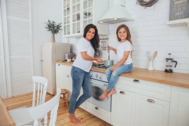 Mutlu bir aile yemeği hazırlıyor. Gülümseyen anne ve kız çocuğu mutfakta yemek pişiriyor ve eğleniyorlar. Ev yapımı sağlıklı yiyecekler. Beyaz İskandinav tarzı iç mekanda küçük bir yardımcı