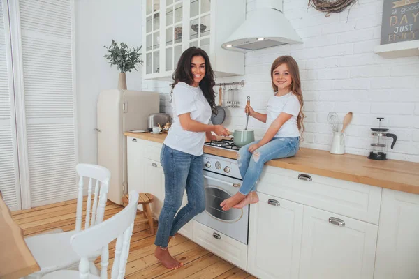 Mutlu bir aile yemeği hazırlıyor. Gülümseyen anne ve kız çocuğu mutfakta yemek pişiriyor ve eğleniyorlar. Ev yapımı sağlıklı yiyecekler. Beyaz İskandinav tarzı iç mekanda küçük bir yardımcı