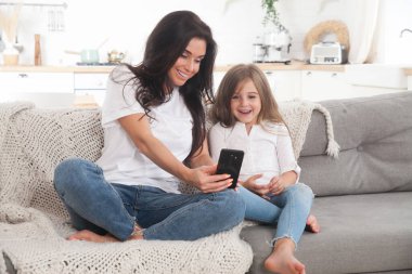 Çekici genç anne ve küçük kız akıllı telefon kullanarak online oyun oynuyorlar, görüntülü sohbet ediyorlar ya da film izliyorlar. Uzaktan eğitim. Karantina ile ilgili video konferansı veya sanal olay