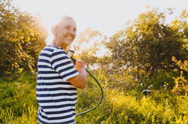 Mutlu yaşlı bir kadın yaz bahçesinde çiçekleri hortumla sularken eğleniyor. Arka planda bir damla su. Çiftçilik, bahçıvanlık, tarım, yaşlı insanlar. Çiftlikte organik sebzeler yetiştiriyor.. 