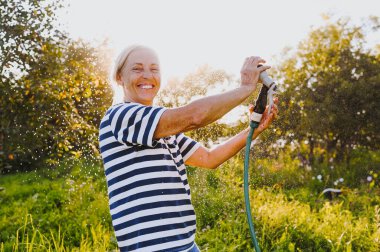 Mutlu yaşlı bir kadın yaz bahçesinde çiçekleri hortumla sularken eğleniyor. Arka planda bir damla su. Çiftçilik, bahçıvanlık, tarım, yaşlı insanlar. Çiftlikte organik sebzeler yetiştiriyor.. 