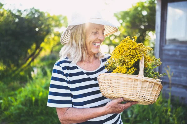 Happy smiling elderly senior woman in straw hat having fun posing in summer garden with flowers in basket. Farming, gardening, agriculture, retired old age people. Growing organic plants on farm.