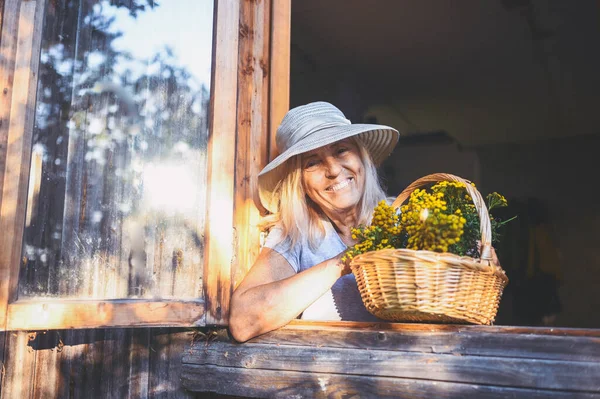 바구니를 모자를 집에서 노부인이 창문을 재미있게 포즈를 취하는 행복하게 미소짓고 — 스톡 사진