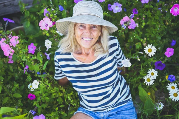 Mutlu gülümseyen yaşlı kadın hasır şapkalı çiçeklerle yaz bahçesinde poz verirken eğleniyor. Çiftçilik, bahçıvanlık, tarım, emekli yaşlılar konsepti. Çiftlikte organik bitkiler yetiştiriyor.. 