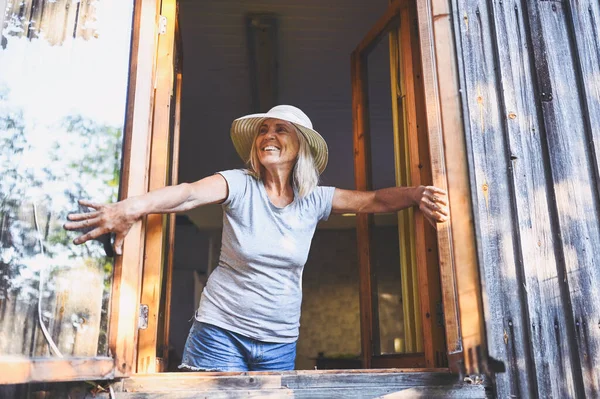 Happy smiling emotional elderly woman having fun posing by open window in rustic old wooden village house in straw hat. Retired old age people concept. Quarantine in the country house
