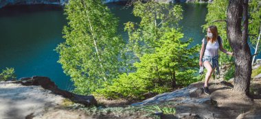 Yeşil ağaçlar ve mavi dağ gölüyle uçuruma doğru yürüyen genç bir kadın Covid 19 önleme için maske takıyor. Sırt çantalı gezgin turist. Corona virüsü ve yerli turizm