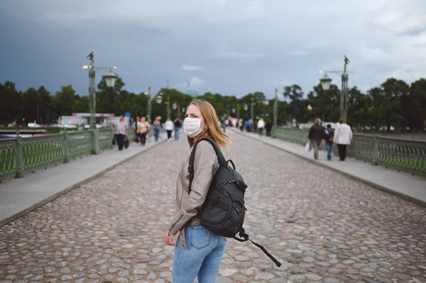 Young blonde woman walking in city street wearing face protective mask for Covid 19 prevention. Caucasian young student or traveler tourist among the crowd with backpack. Corona virus concept.