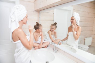 Dişlerini fırçalayan ve birlikte vakit geçiren mutlu bir aile. Anne ve kızı beyaz banyoda öpüşüp sarılıyorlar. Kafalarında ve vücutlarında havlular olan kadın ve kız. 