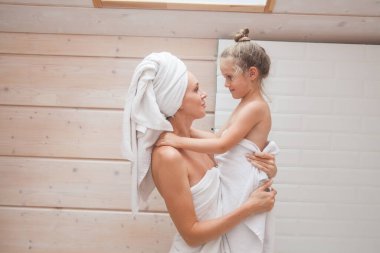 Mutlu sevgi dolu aile birlikte vakit geçiriyor. Anne ve kızı beyaz banyoda öpüşüp sarılıyorlar. Kafalarında ve vücutlarında havlular olan kadın ve kız. 