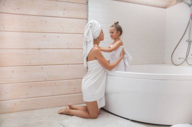 Mutlu sevgi dolu aile birlikte vakit geçiriyor. Anne ve kızı beyaz banyoda öpüşüp sarılıyorlar. Kafalarında ve vücutlarında havlular olan kadın ve kız. 