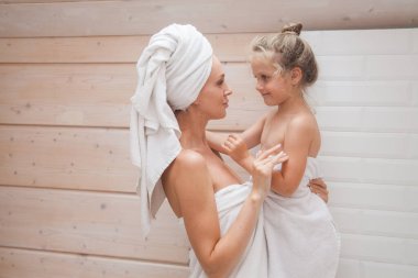 Mutlu sevgi dolu aile birlikte vakit geçiriyor. Anne ve kızı beyaz banyoda öpüşüp sarılıyorlar. Kafalarında ve vücutlarında havlular olan kadın ve kız. 