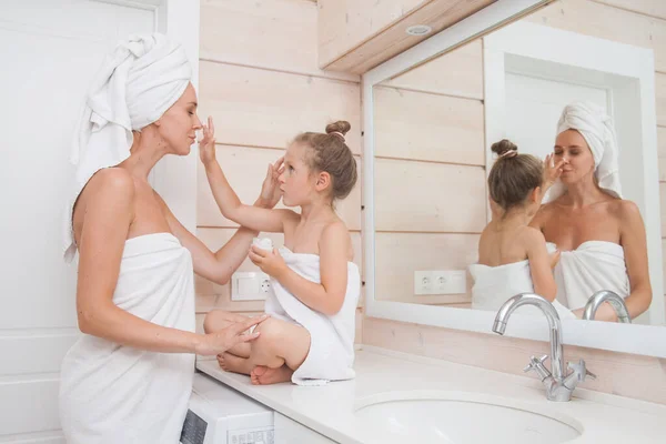 Happy loving family take care of the skin and apply moisturizer on face. Mother and her daughter child girl having fun in white bathroom. Woman and girl with towels on their heads and bodies
