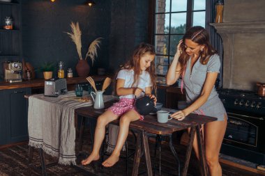 Mutlu bir aile yemeği hazırlıyor. Gülümseyen anne ve kız çocuğu mutfakta yemek pişiriyor ve eğleniyorlar. Ev yapımı sağlıklı yiyecekler. Evdeki karanlık bir mutfakta küçük bir yardımcı..