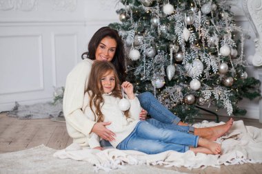 Yeni yıl arifesinde mutlu bir aile. Süslü süveter ve battaniye içinde güzel bir anne ve tatlı bir kız çocuğu süslü bir Noel ağacının altında eğleniyor ve sarılıyorlar. Şenlikli ev klasiği iç mimarisi.