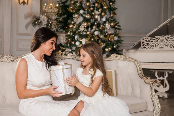 Merry Christmas and Happy Holidays. Cheerful mom and her cute daughter girl exchanging gifts in white classic interior against the background of a piano and a decorated Christmas tree. New Year 2021